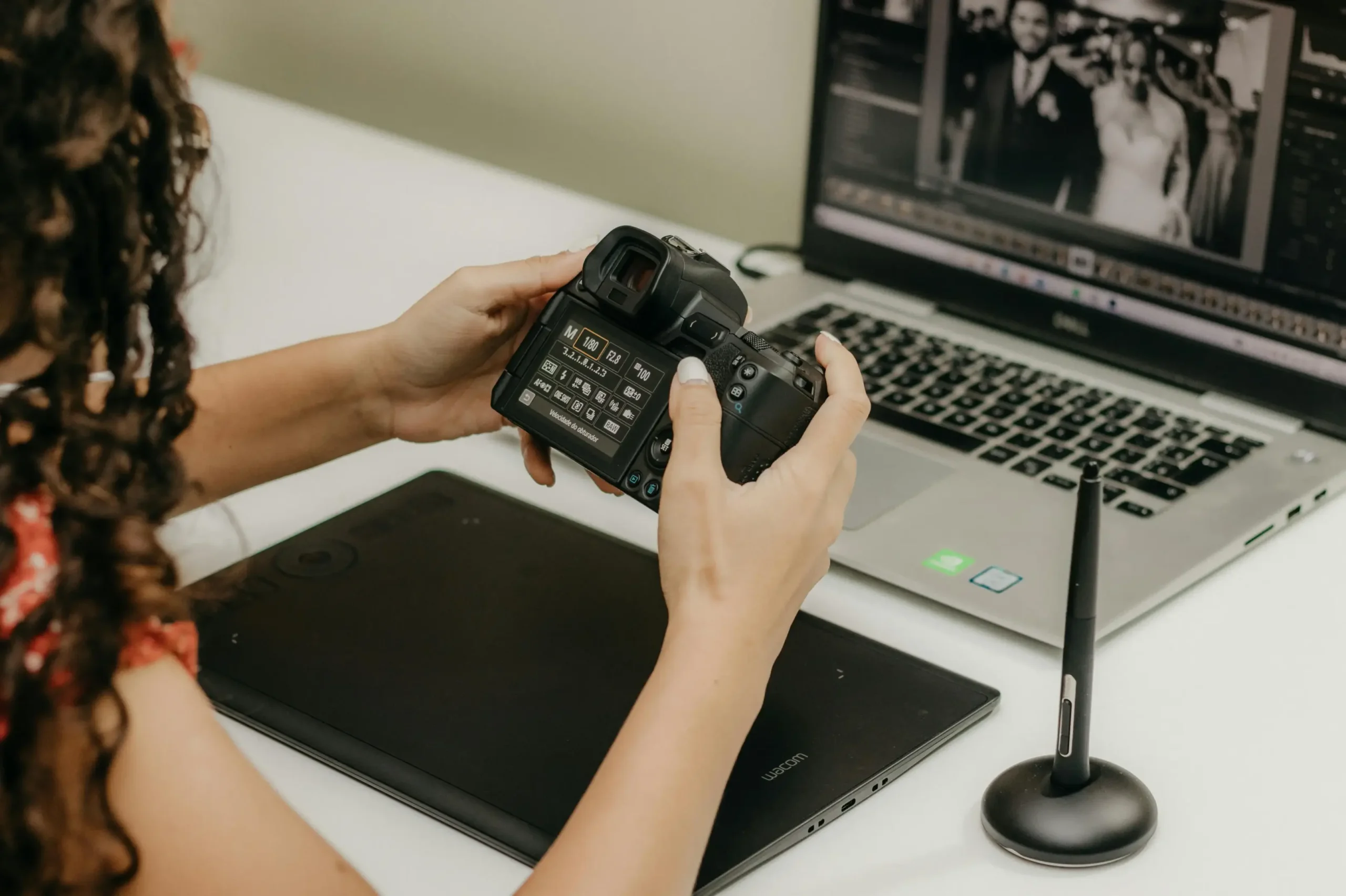 A person with long curly hair holds a professional camera while sitting at a desk equipped with a laptop and a digital drawing tablet. The laptop screen displays a black-and-white wedding photograph being edited, highlighting a creative workflow between photography and digital post-production.