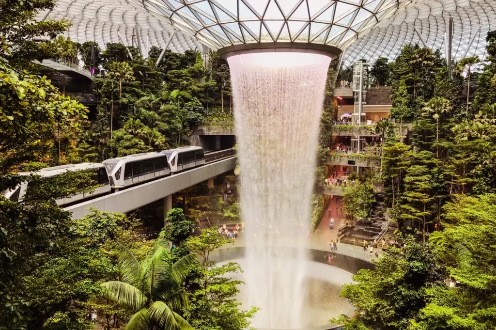 An indoor waterfall cascades down into a pool surrounded by lush greenery and a monorail passing by under a domed glass ceiling.