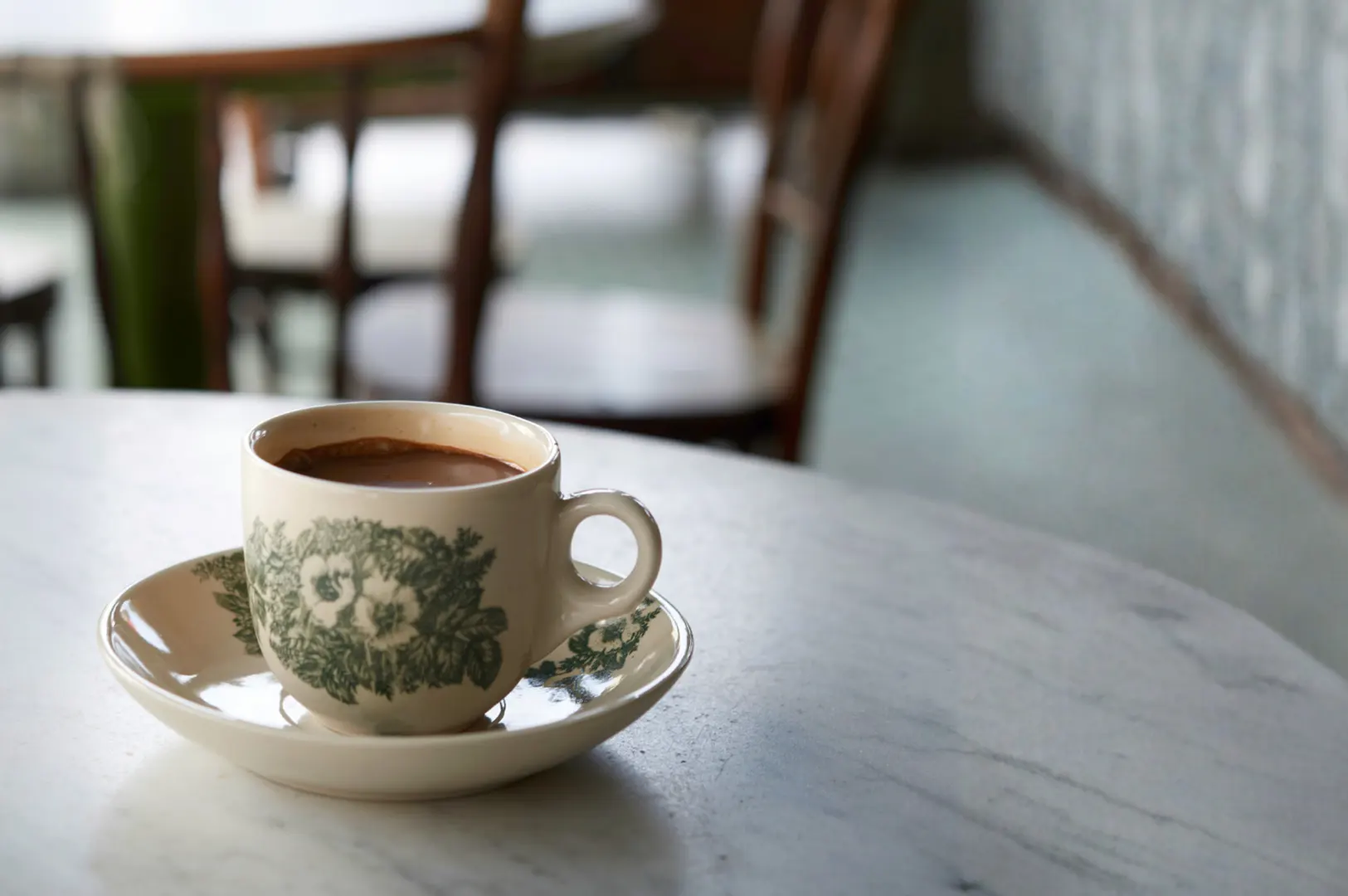 A cup of coffee with a floral pattern sits on a saucer on a marble table. In the background, wooden chairs are visible.