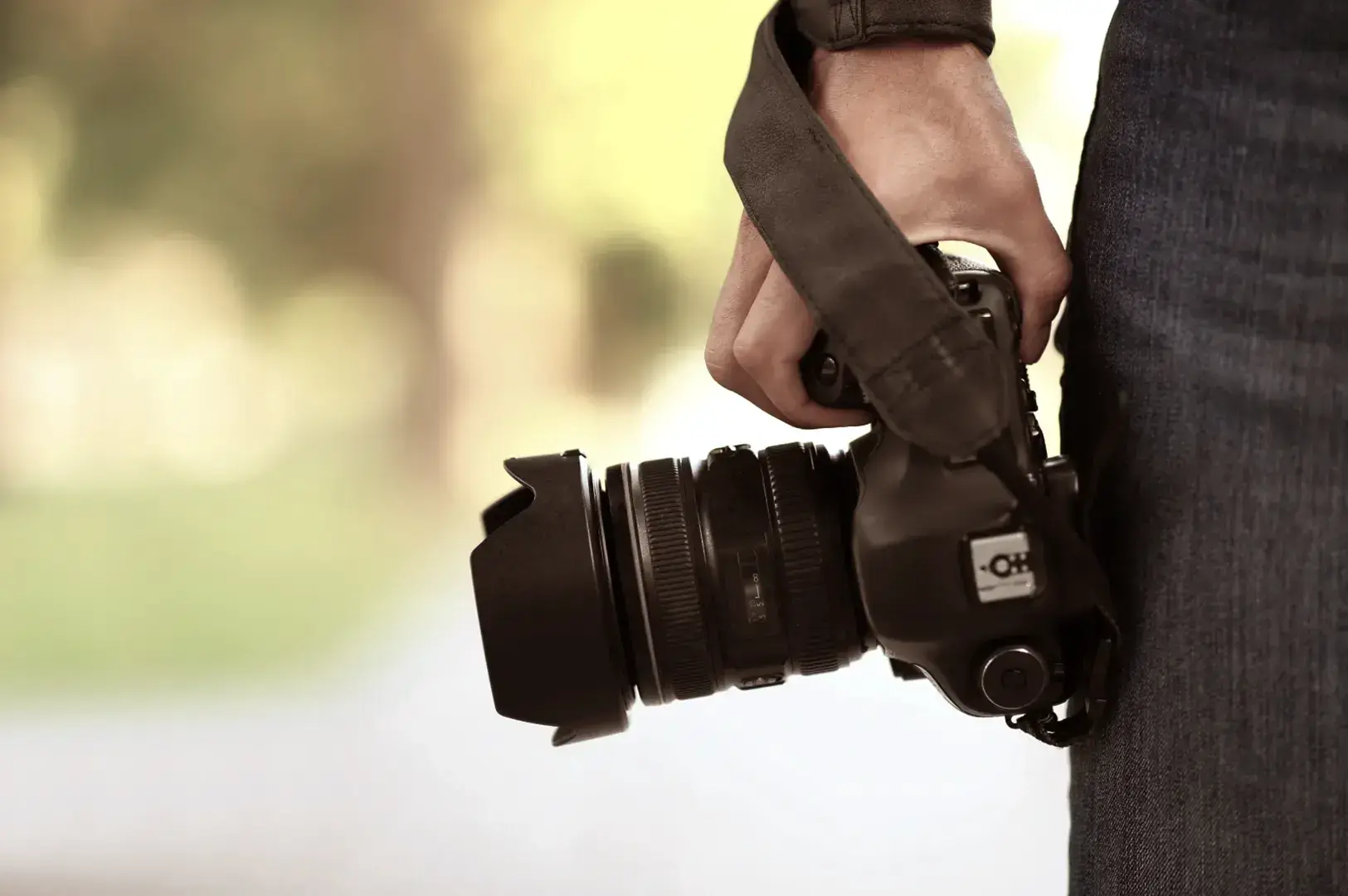 A person holds a black DSLR camera with a lens hood attached, wearing blue jeans.