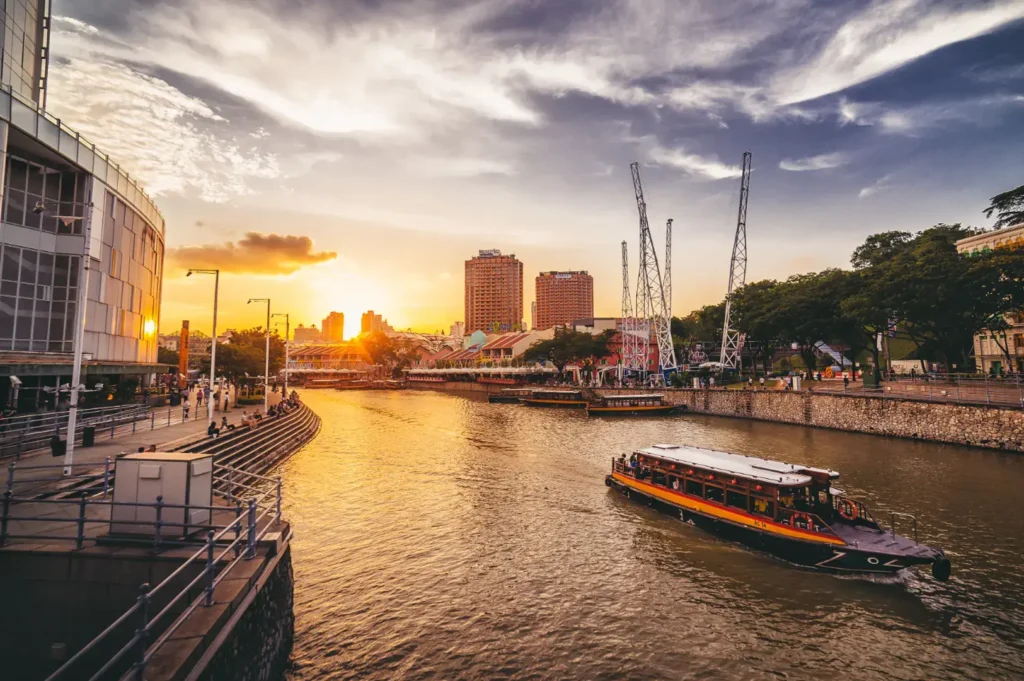 A riverboat navigates a river lined with buildings and trees during a sunset with dramatic clouds.