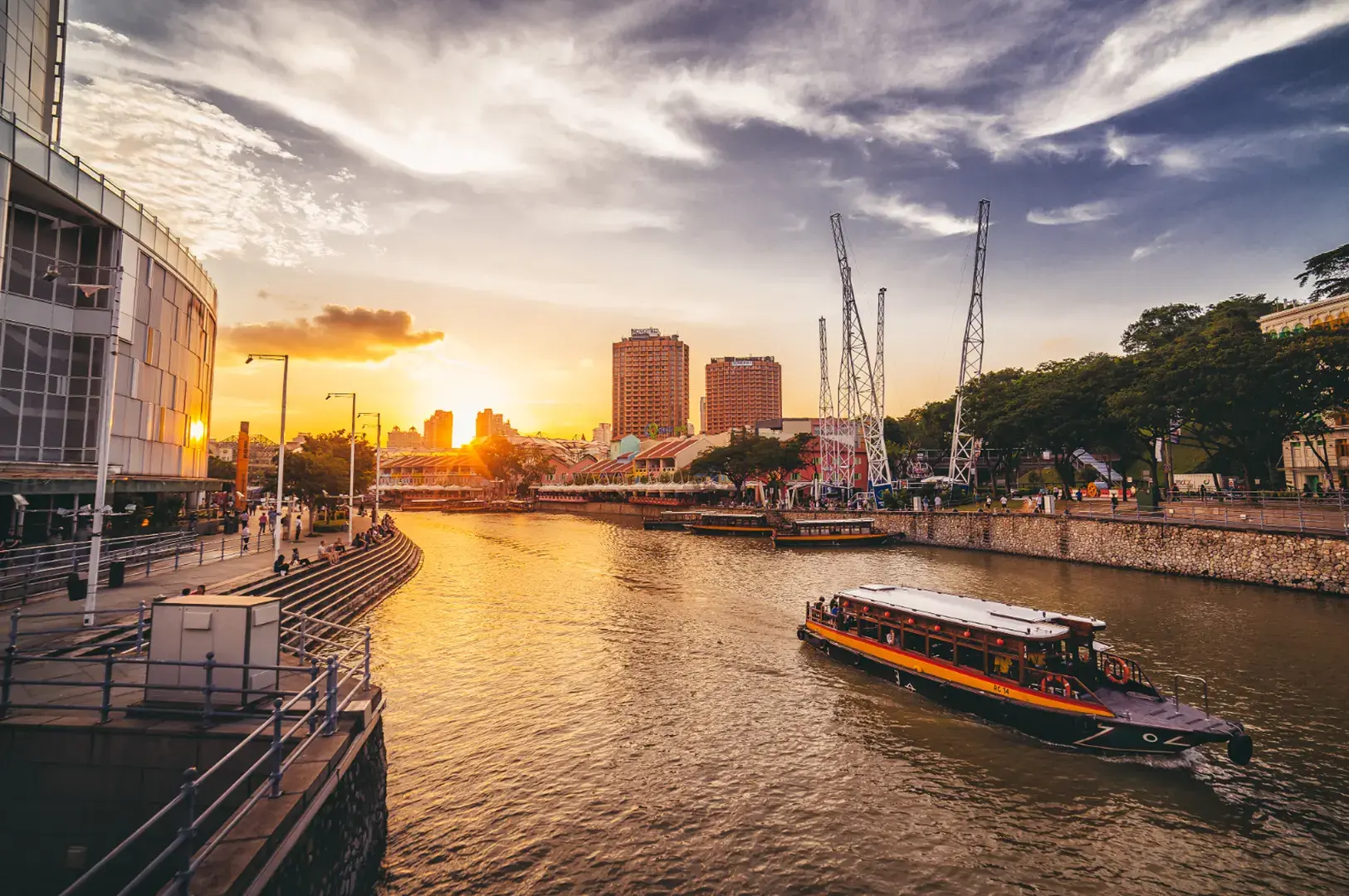 A riverboat navigates a river lined with buildings and trees during a sunset with dramatic clouds.