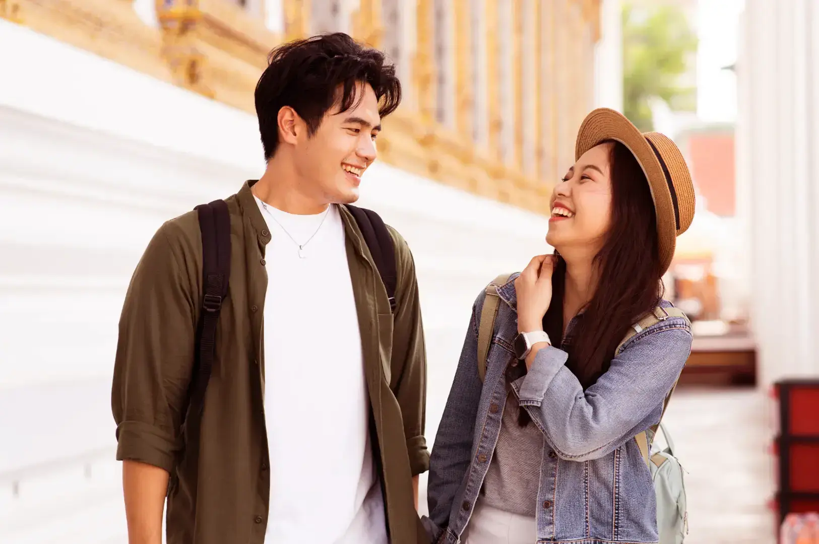 A smiling couple holding hands and looking at each other while wearing backpacks, with a temple in the background.