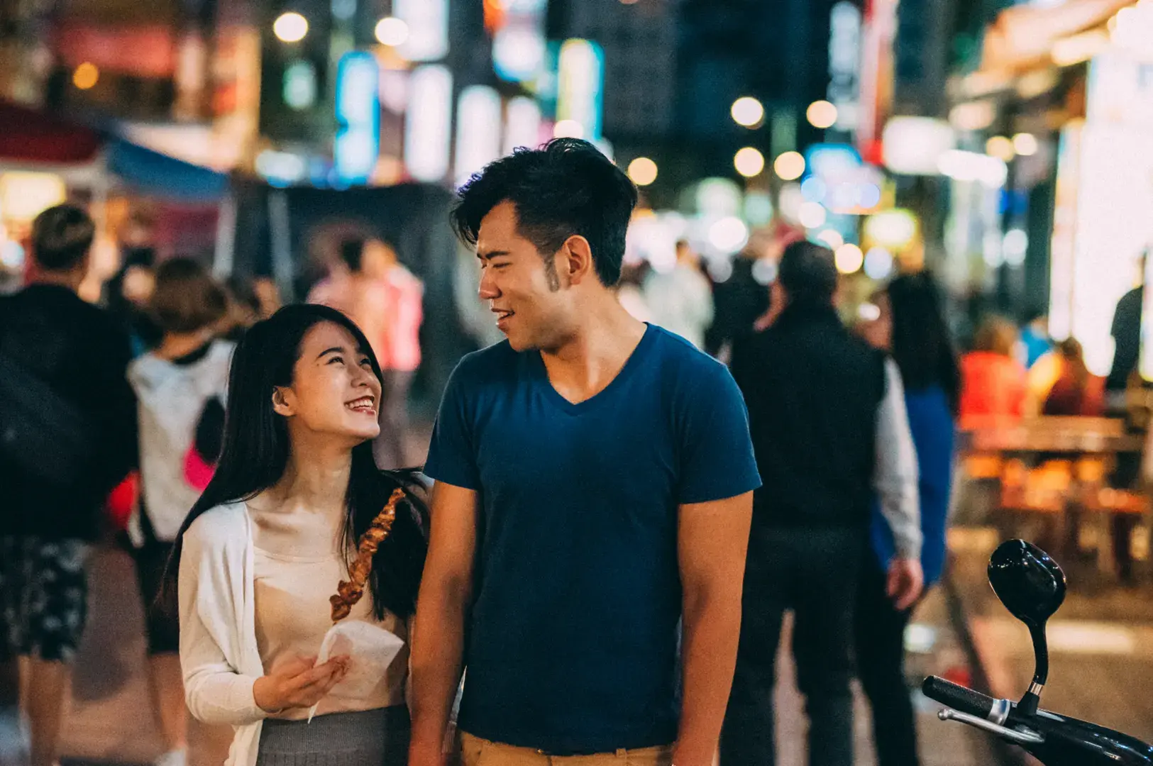 A couple smiles at each other while walking down a busy, brightly lit street at night, with food in hand.