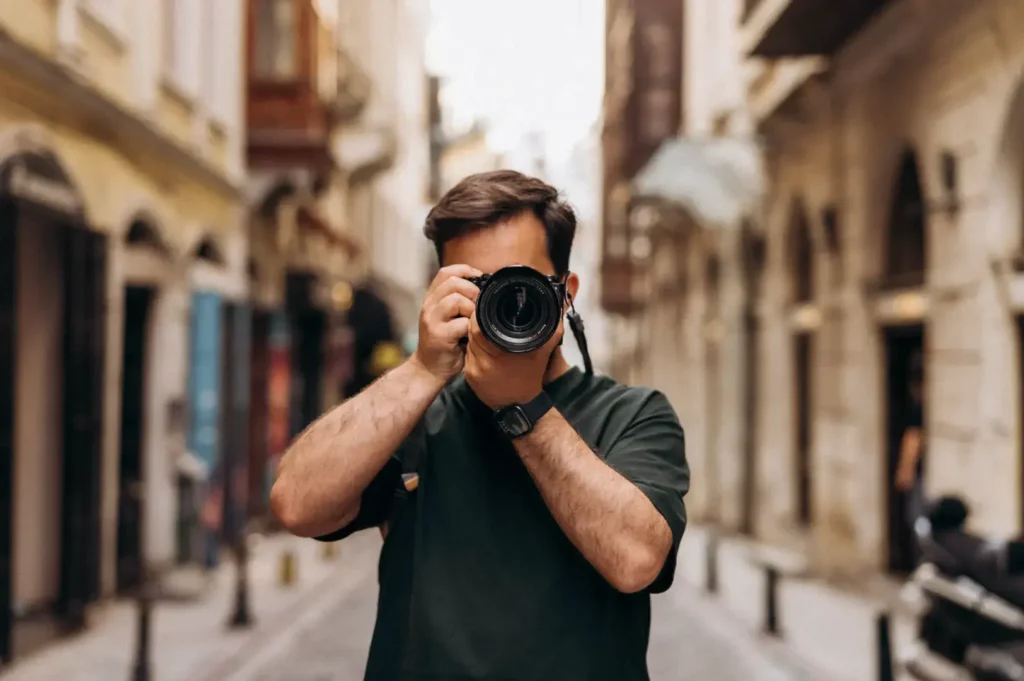 A person in a dark green t-shirt holds a camera up to their face, framing a shot down a narrow street lined with buildings.