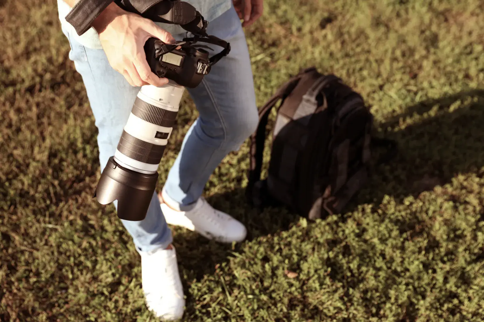 A person in jeans and white sneakers holds a professional camera with a long lens. A black backpack rests on the grass beside them.
