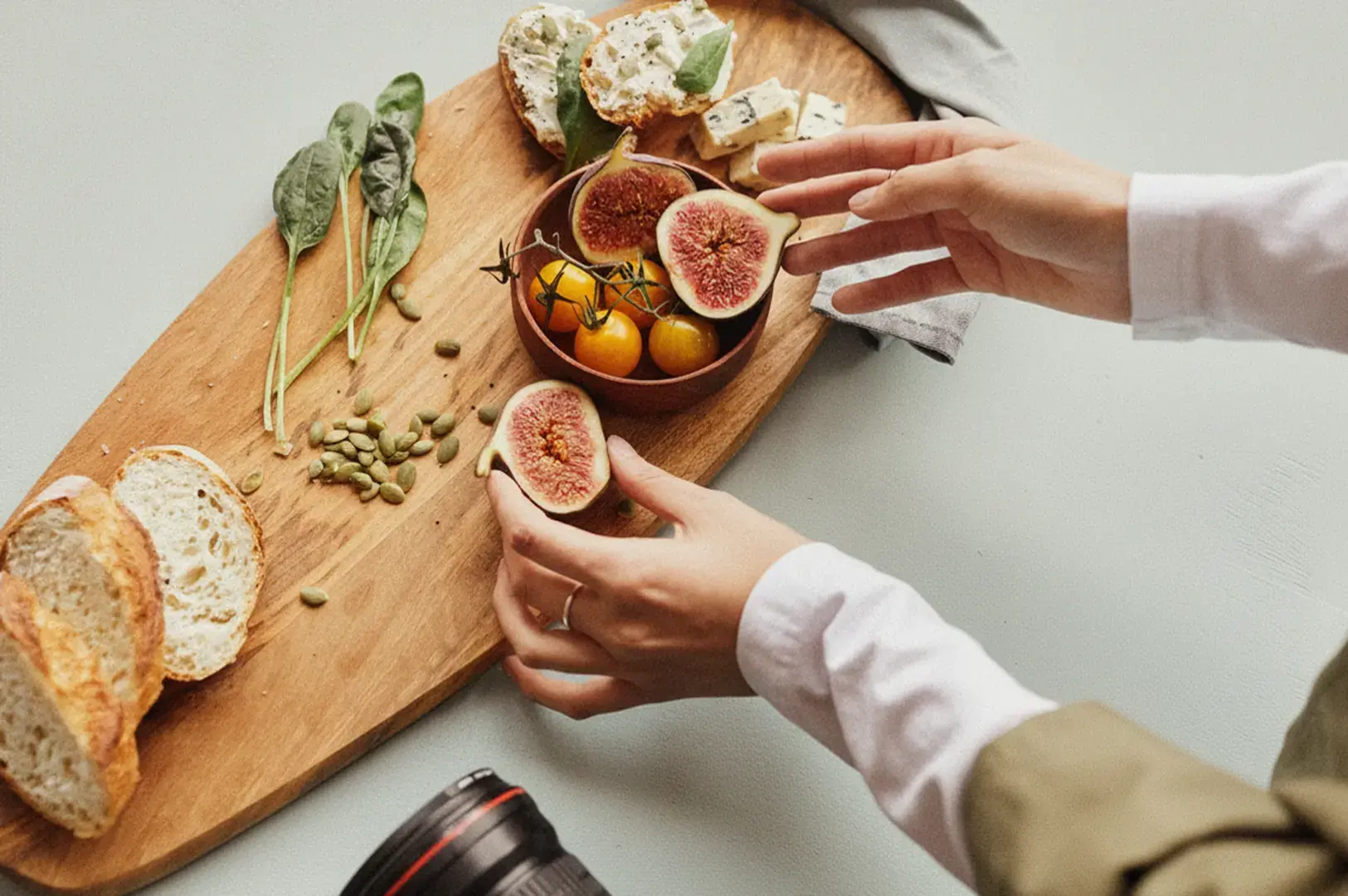 A person arranging food on a board beside a camera on a clean tabletop.