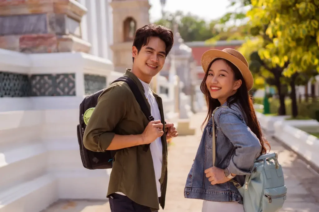 A man and a woman smiling at the camera while wearing backpacks and standing outdoors next to a white structure with intricate green patterns.