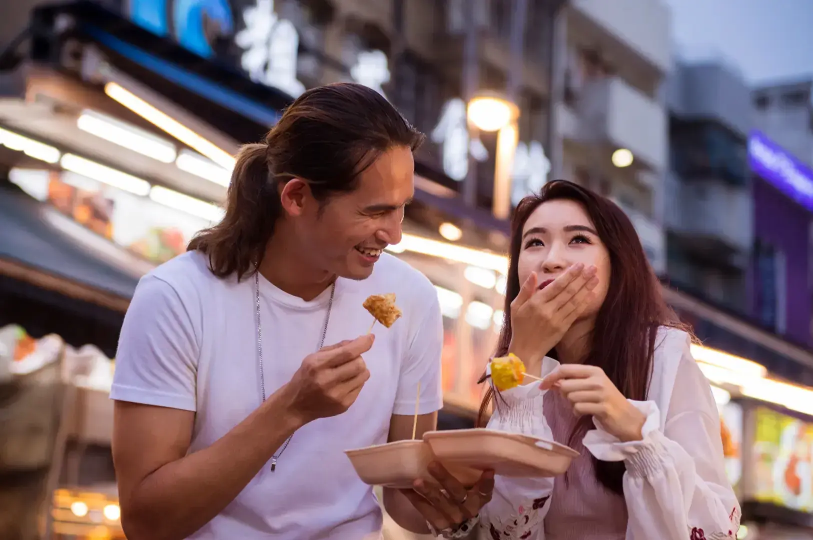 A couple enjoys street food at night with glowing market stalls in the background. The man holds a skewer of fried food, while the woman covers her mouth while eating.