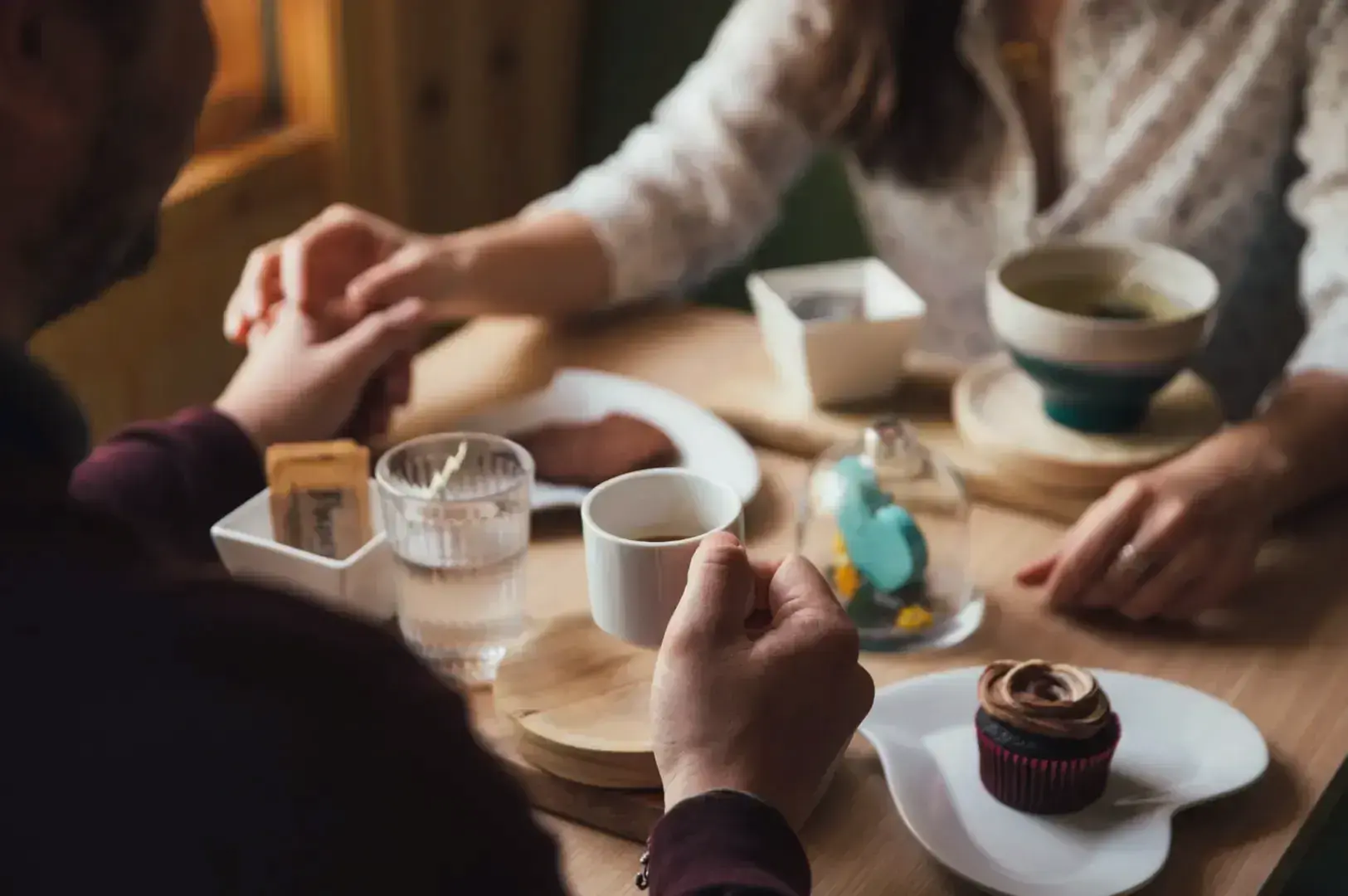 A couple holds hands across a table laden with coffee, a cupcake, and a cookie.