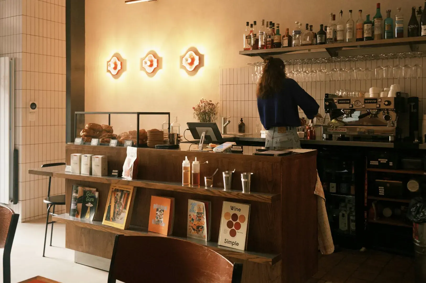 Warm-lit cafe counter with staff working at espresso machine, retail shelving with packaged goods, and circular wall lights.