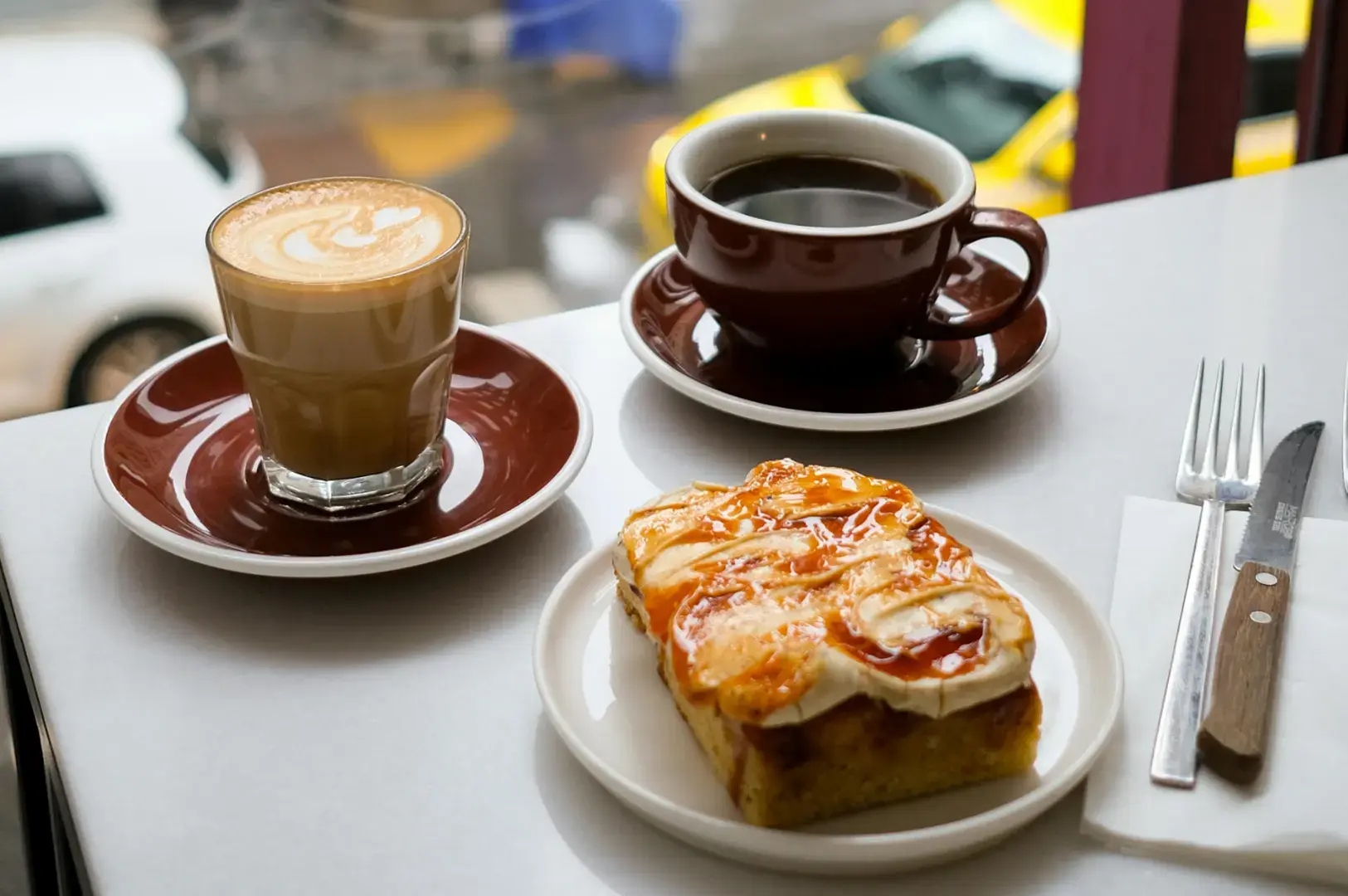 Latte with latte art and black coffee in brown cups on saucers beside a cheese-topped pastry on a white plate with fork and knife.