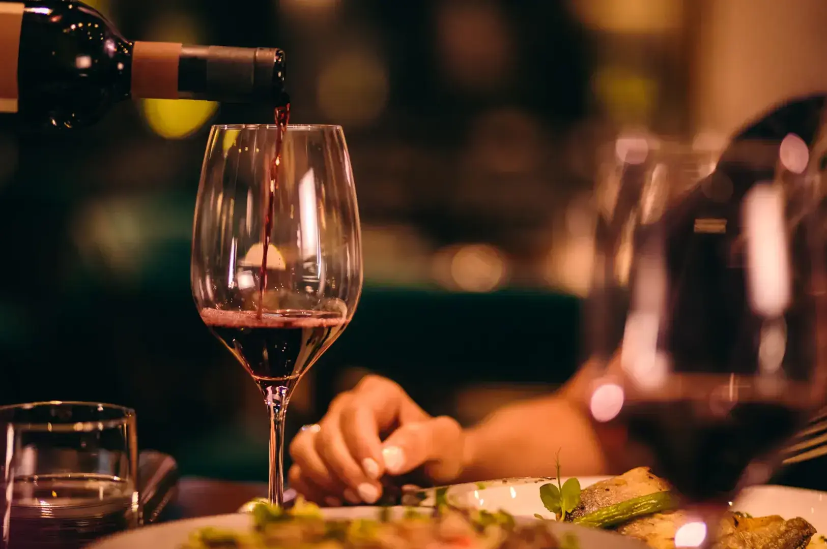 Red wine being poured from a bottle into a glass on a table with food and other glasses.