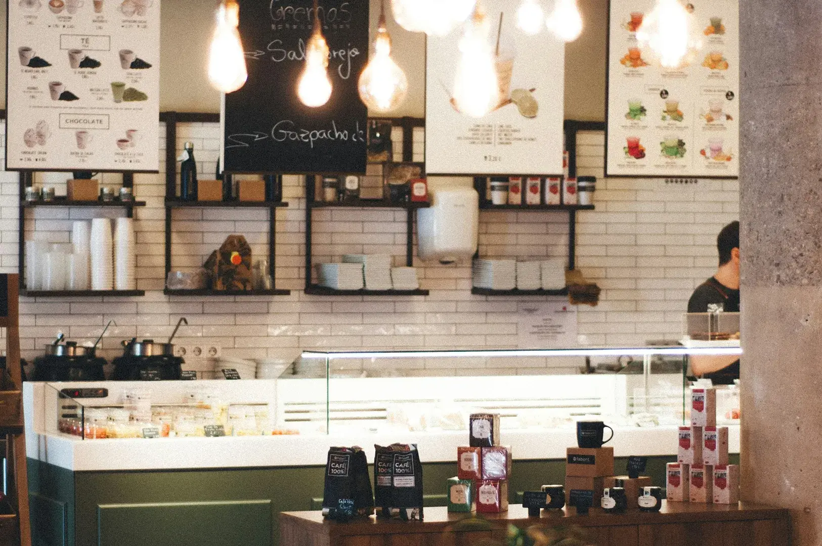 Cafe counter with white subway tile backsplash, hanging Edison bulb lights, chalkboard menu, open shelving with coffee products, and barista working behind green-paneled service counter.