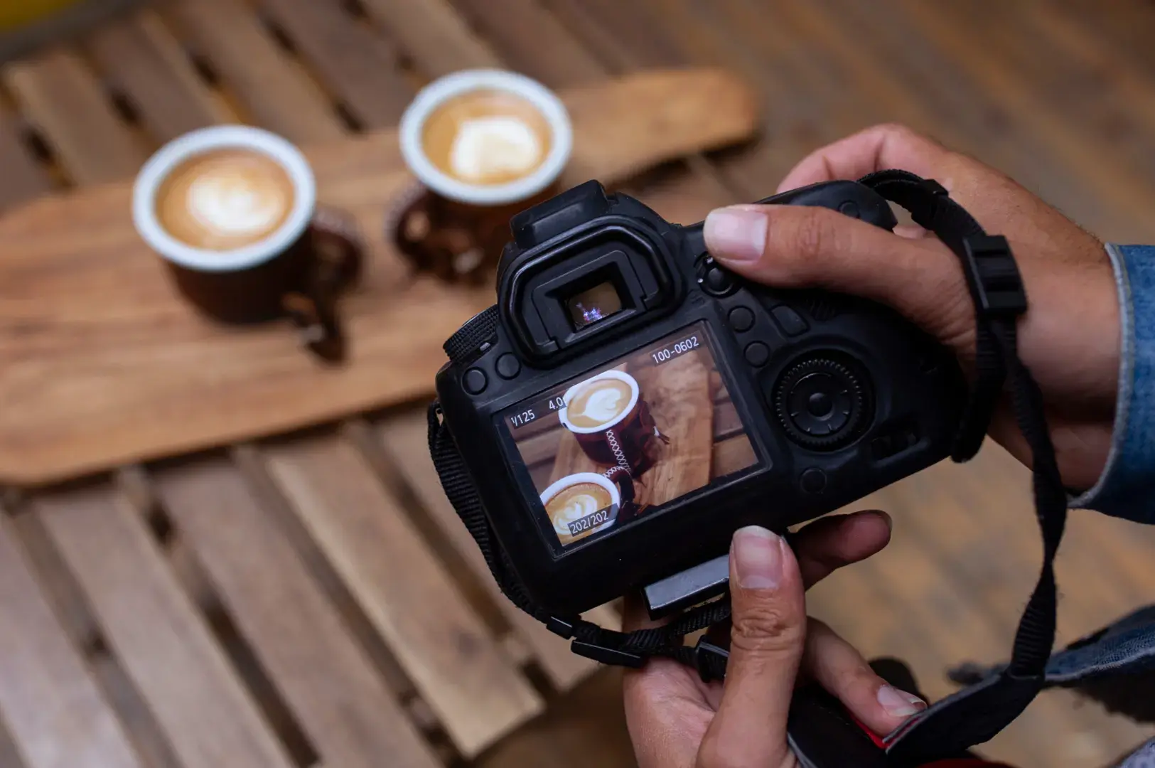 Hands holding DSLR camera reviewing latte art photo on screen with two coffee cups on wooden table in background.