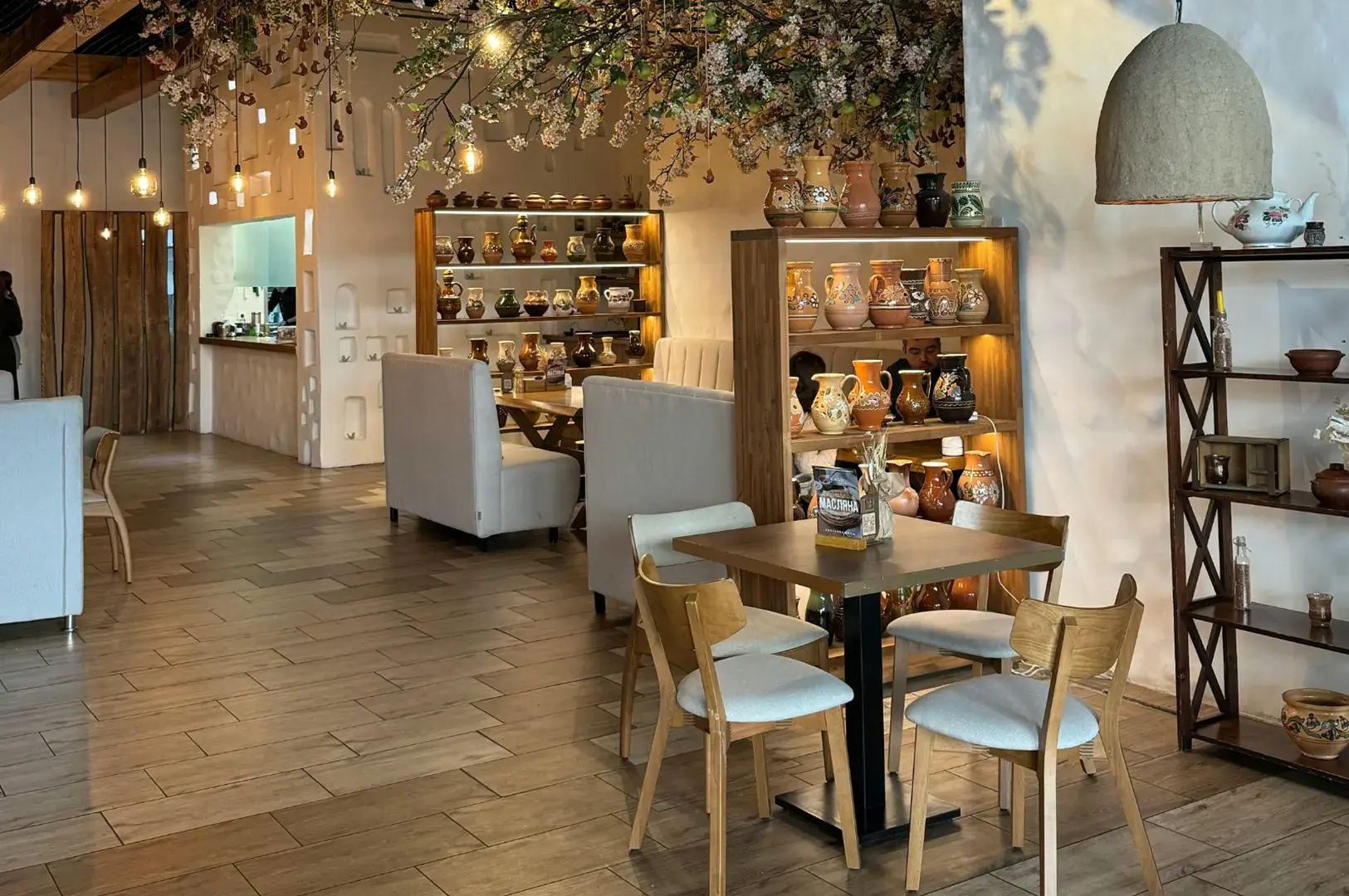Cozy cafe with warm lighting, hanging floral installation, wooden shelving with pottery display, and small dining table with cream chairs.