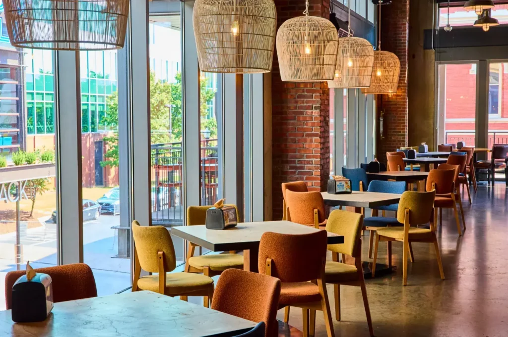 Bright cafe interior with colorful chairs, woven pendant lights, exposed brick wall, and floor-to-ceiling windows overlooking outdoor terrace.