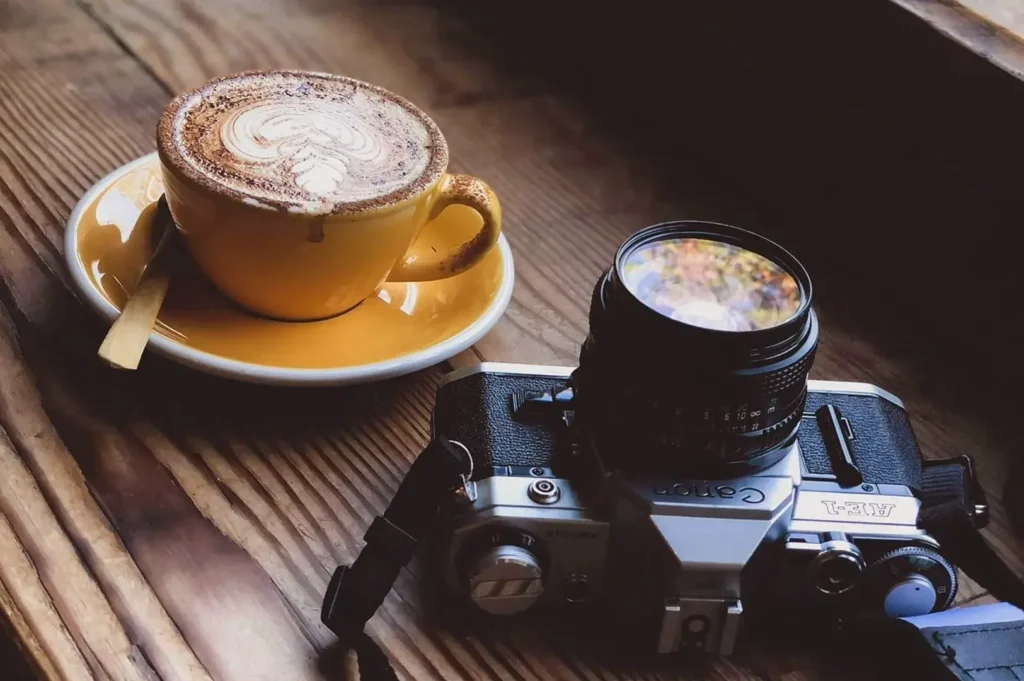 Cappuccino with latte art on yellow saucer beside vintage film camera with lens on wooden slatted table in natural window light.