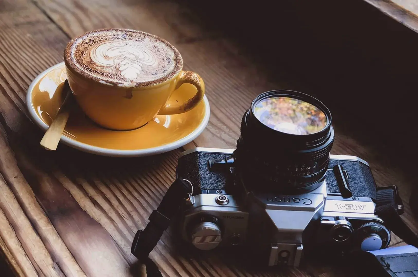 Cappuccino with latte art on yellow saucer beside vintage film camera with lens on wooden slatted table in natural window light.