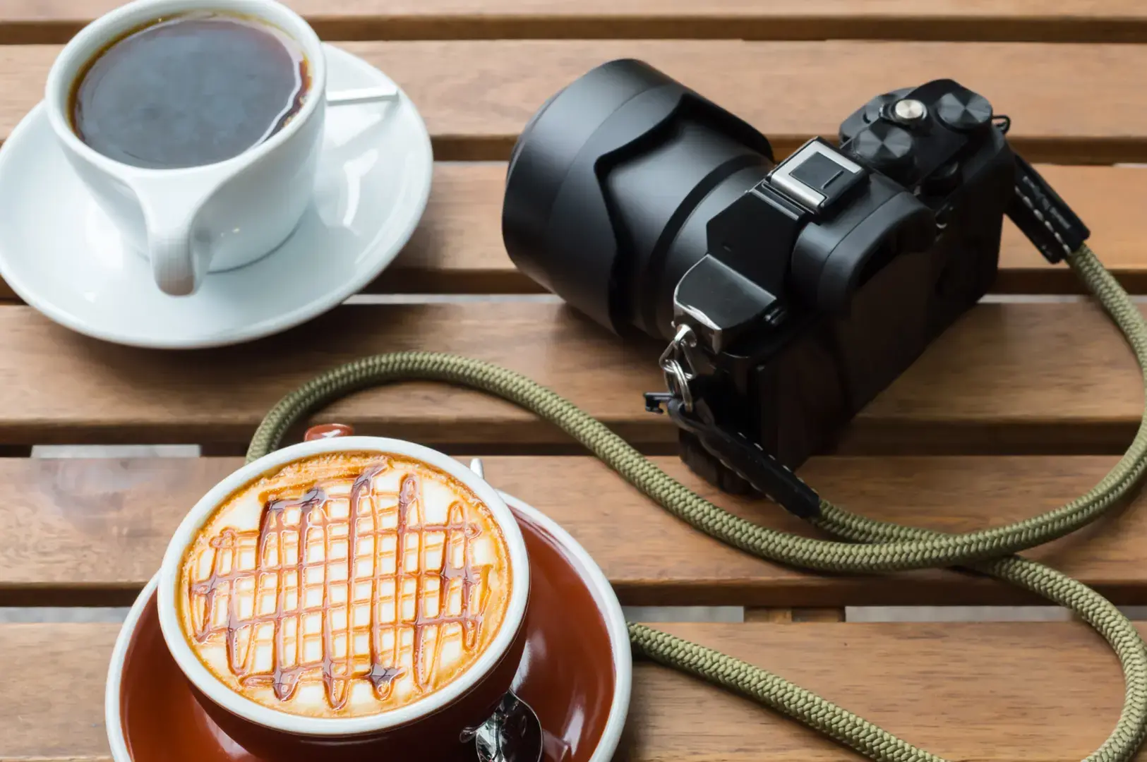 DSLR camera with green strap beside caramel latte and black coffee on wooden slatted table.