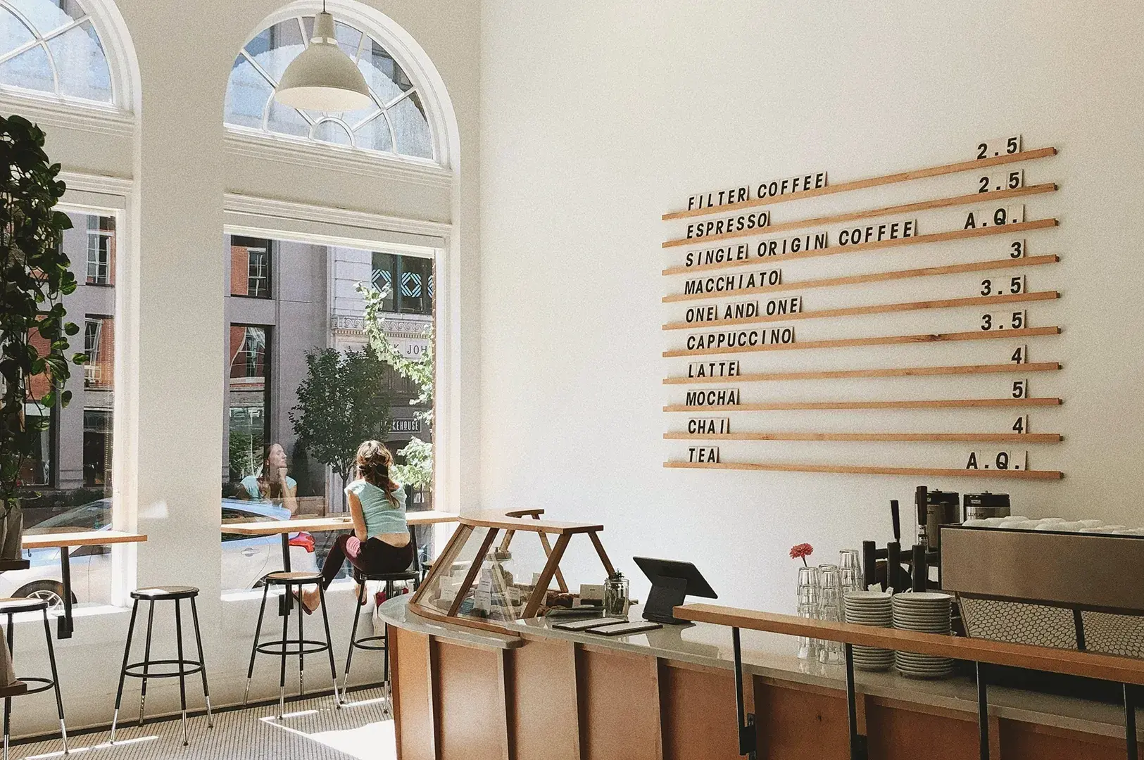 Minimalist cafe interior with wooden menu board displaying coffee prices, arched windows with natural light, wooden counter with espresso machine, and bar seating.
