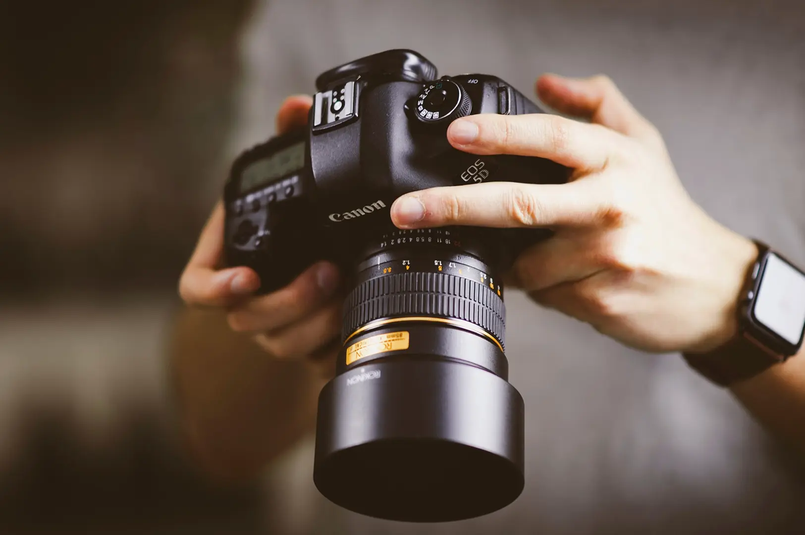 Hands holding Canon DSLR camera with telephoto lens, photographer reviewing settings on camera body in natural light.