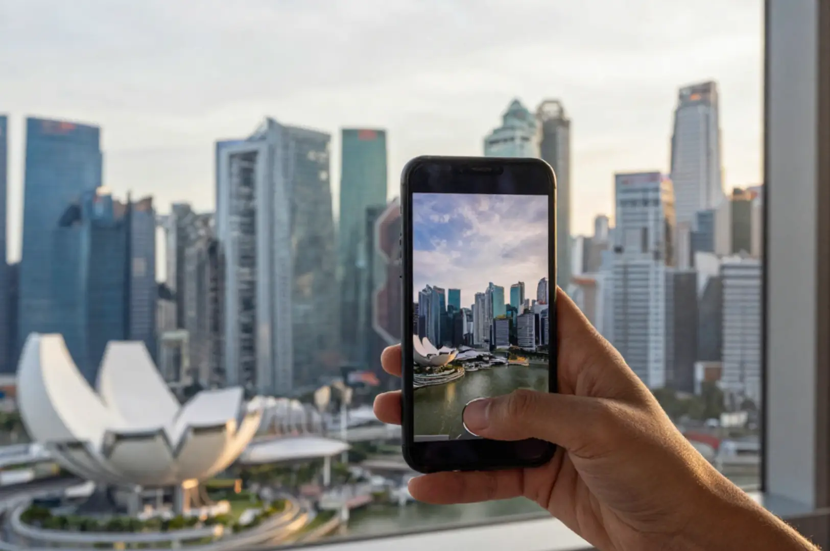 A hand holding a smartphone in front of Singapore’s skyline, with Marina Bay visible in the background.