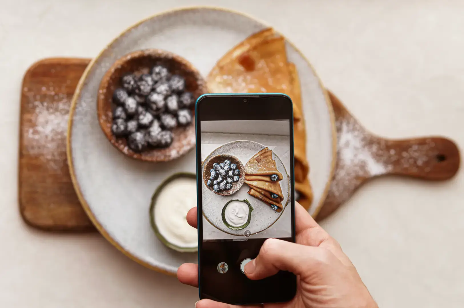 A hand holding a smartphone photographs a plated dessert with powdered sugar, a bowl of dark berries, and small cream and cucumber slices on a wooden serving board.