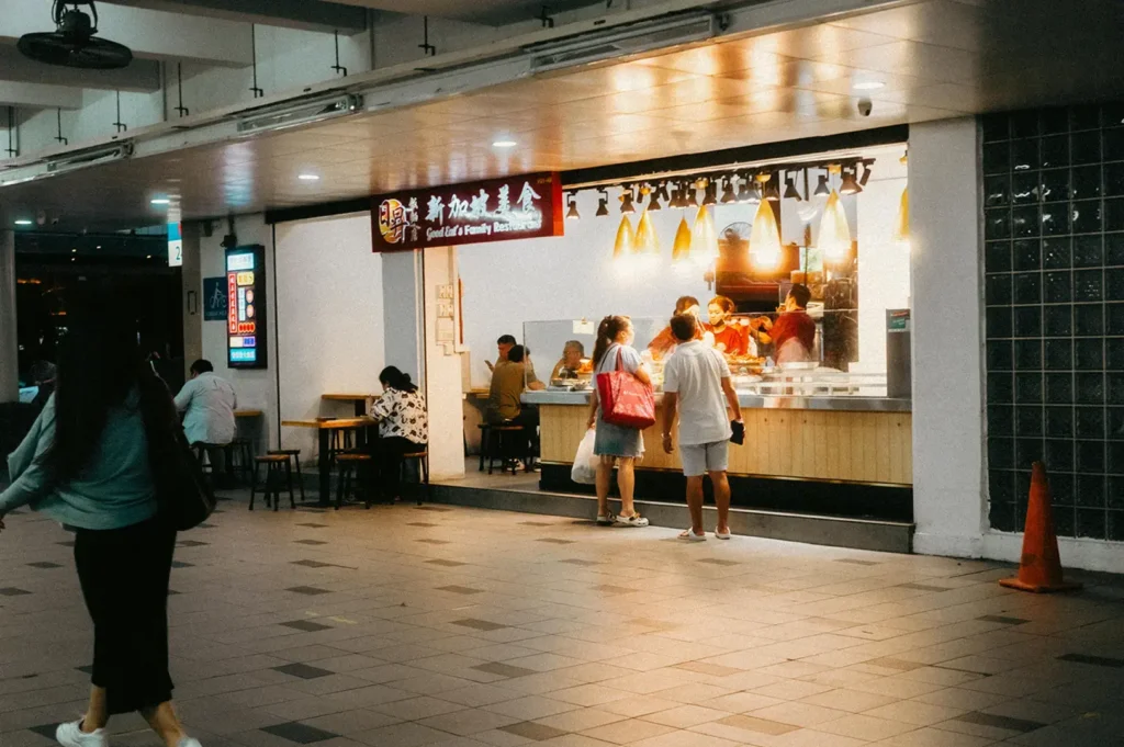 A food stall counter with diners standing nearby in a covered walkway.