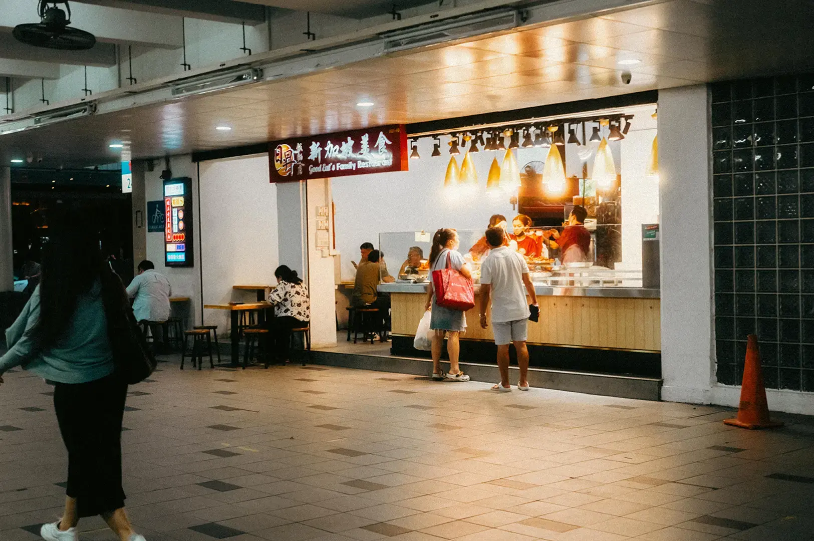 A food stall counter with diners standing nearby in a covered walkway.