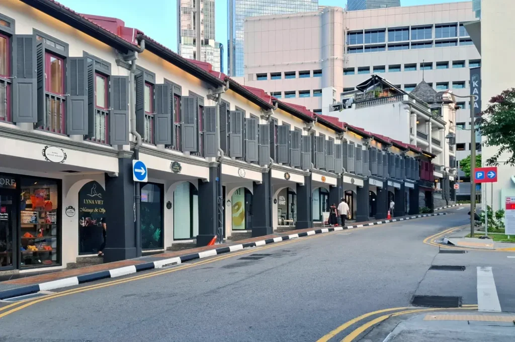 A row of traditional shophouses with gray shutters lines a street with modern buildings in the background.
