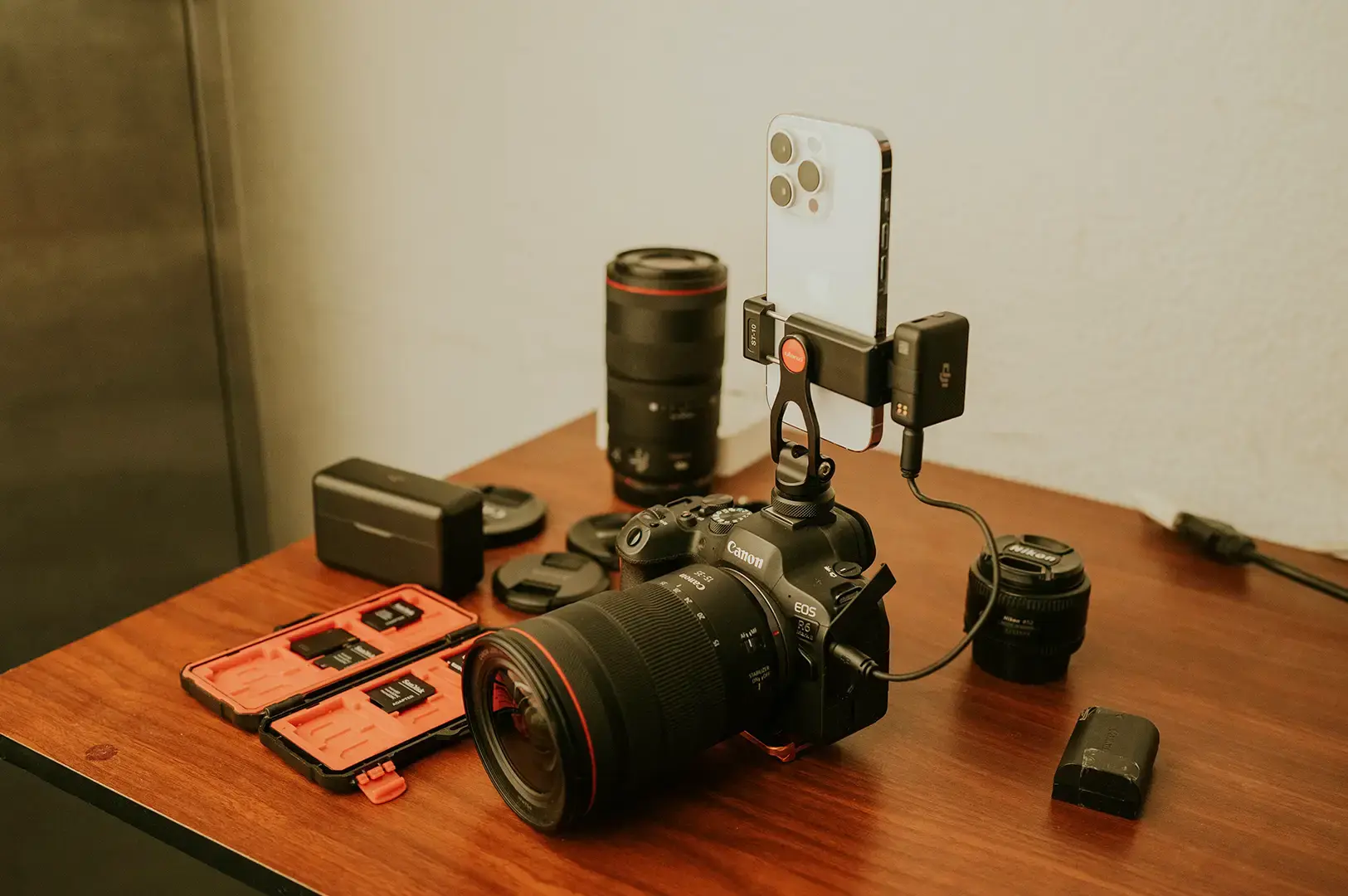 A camera with a large lens, lenses, batteries, and a smartphone arranged on a wooden table.