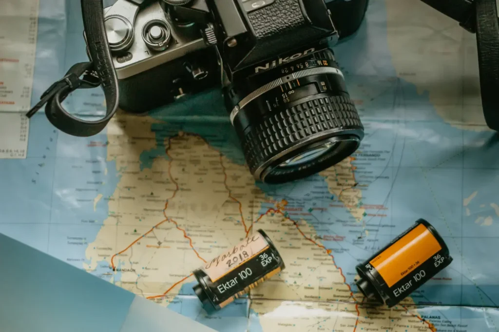 Black vintage camera and film canisters on top of a folded world map, with a small landscape photo showing a person in a red jacket standing on a mountain overlook.