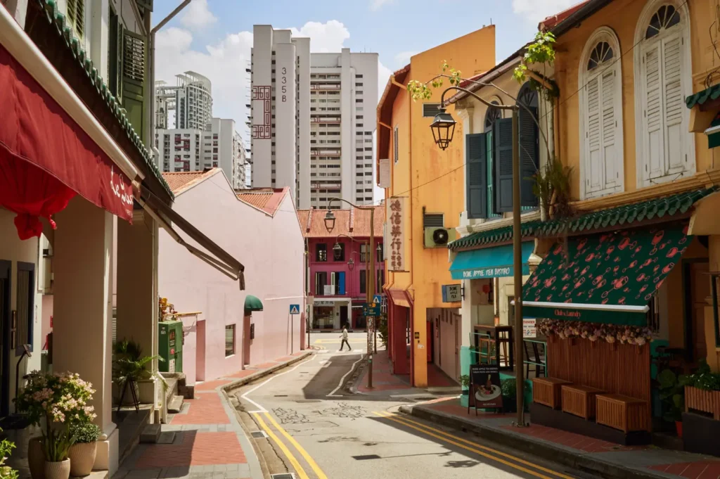 Narrow street in an urban heritage district with red-brick shopfronts and awnings lining both sides, street lamps and planters, and tall apartment buildings in the background under a blue sky with clouds.