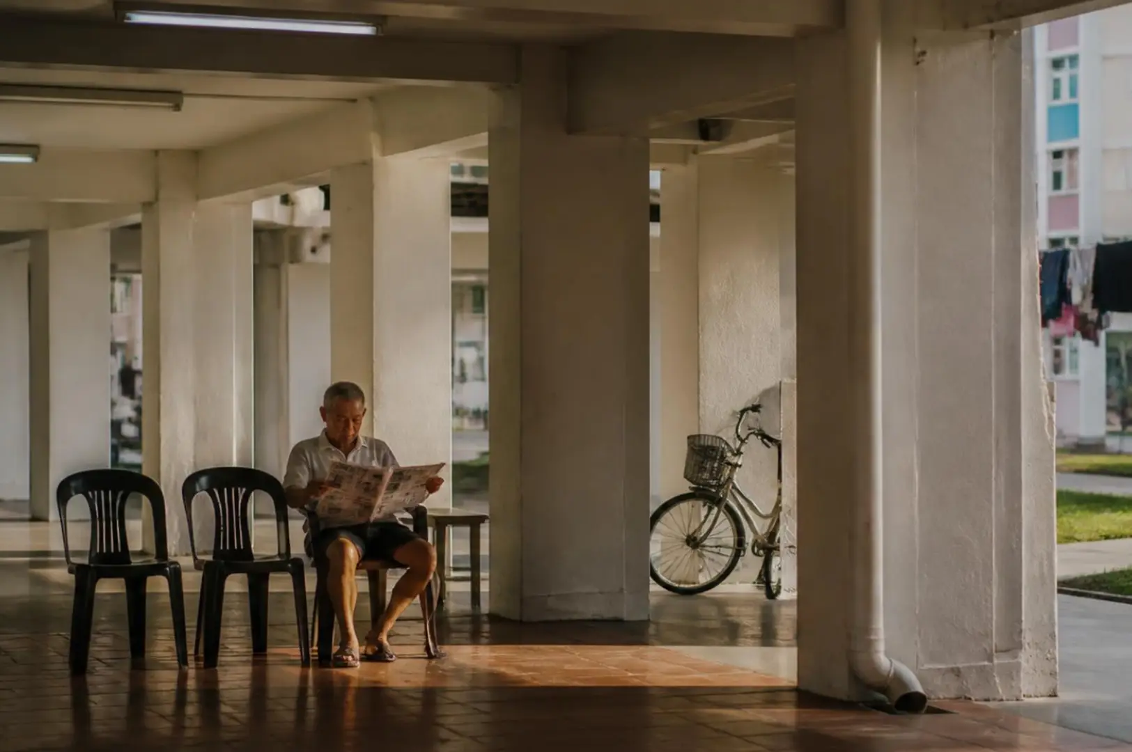 Under a covered concrete walkway, a person sits on the floor reading a paper, surrounded by empty plastic chairs; a bicycle leans against a column near the right side.