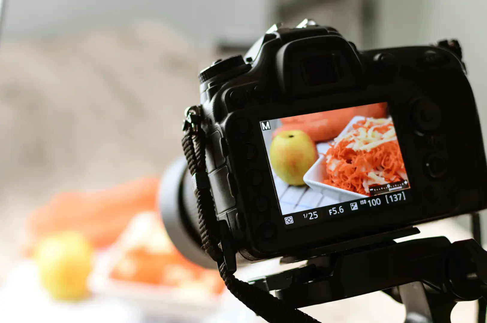 Digital camera mounted on a tripod and focused on a plate of food, with apples and a blurred dining setup in the background.