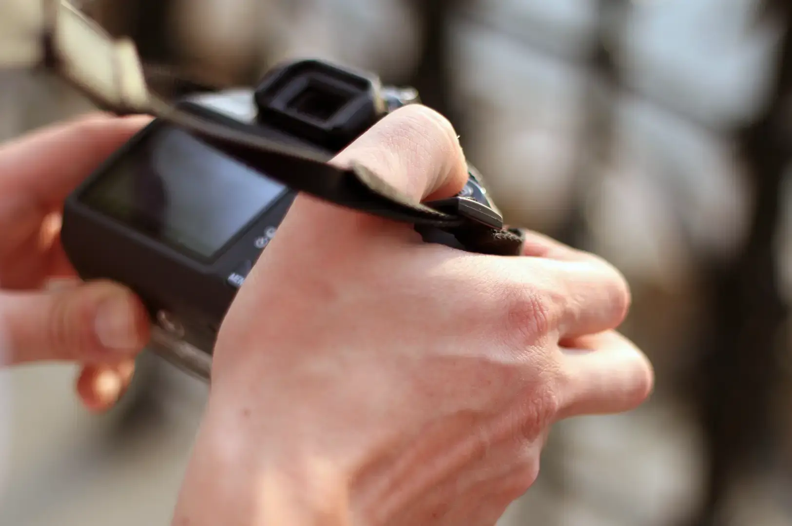 Close-up of a person holding a digital camera with both hands, adjusting the controls while viewing the camera screen outdoors.