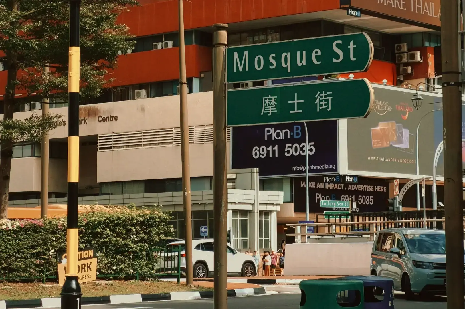 Street scene in front of a large office building with orange awnings and a green street sign reading “Mosque St” alongside a smaller sign, cars parked along the curb.
