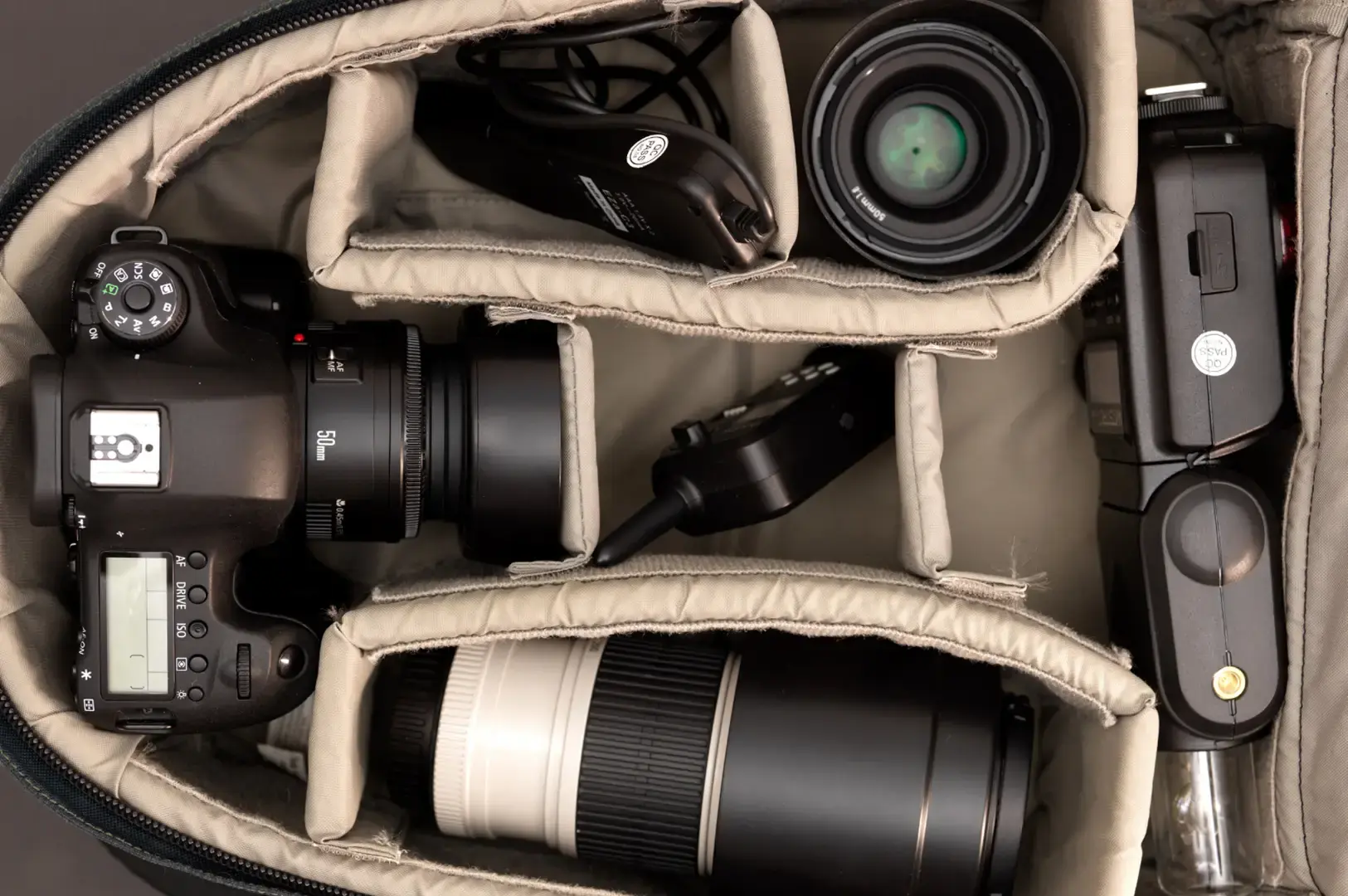 Open camera bag lined with beige fabric showing a DSLR camera, several lenses, a green-tinted viewfinder lens, and accessories neatly arranged in compartments.