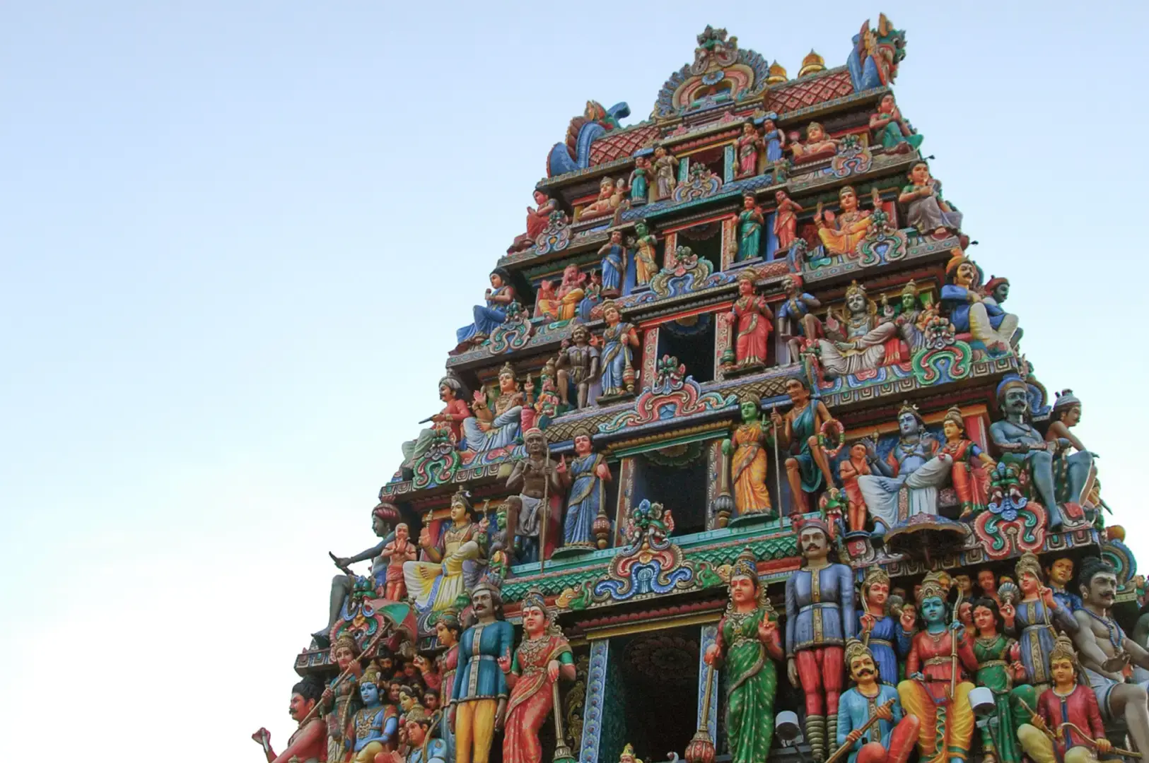Tall temple gopuram covered with colorful painted figures and ornate carvings, viewed from below against a clear blue sky, with a dark entrance opening at the base.