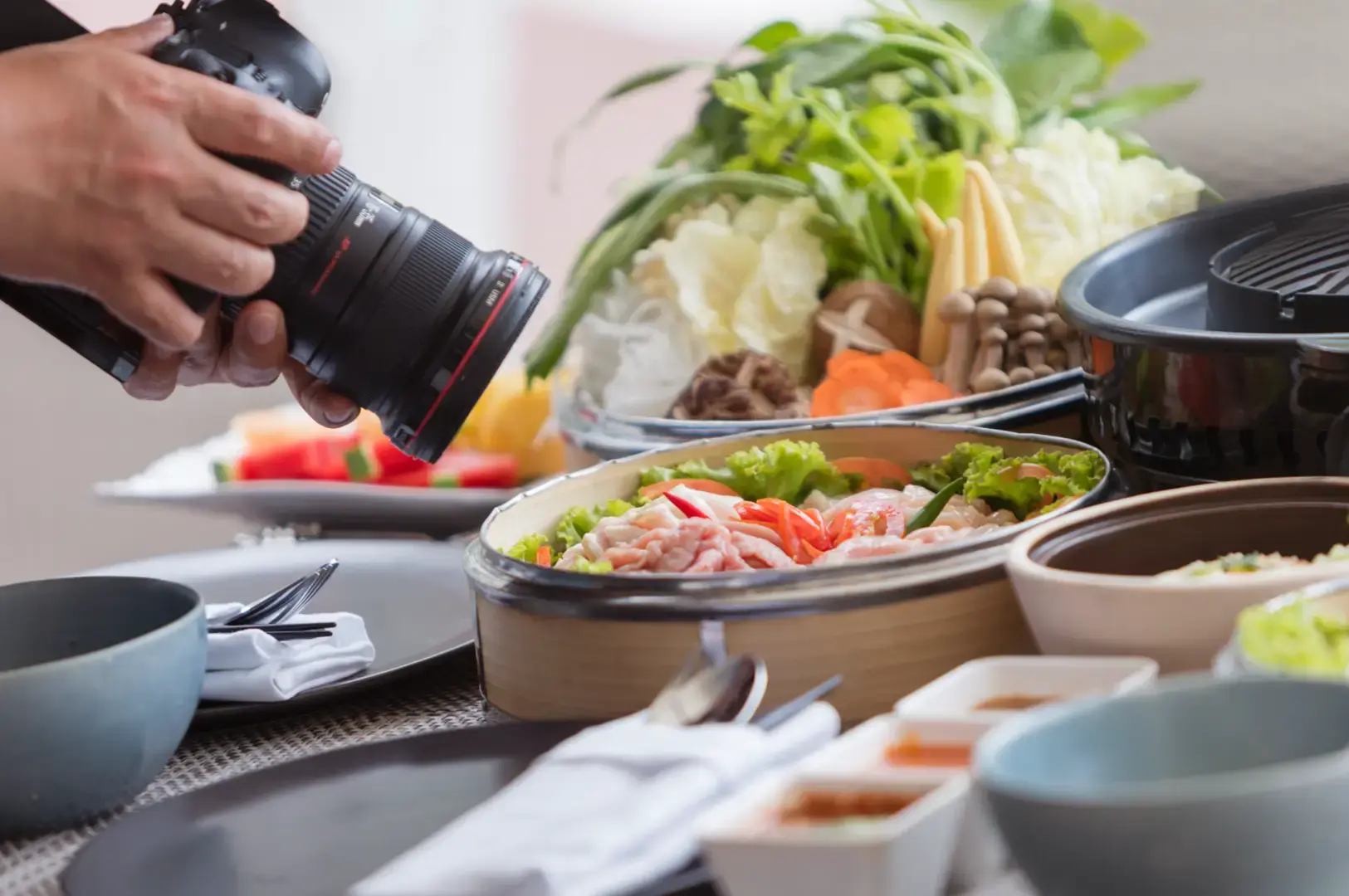 Photographer using a DSLR camera to capture a tabletop meal with seafood, leafy vegetables, mushrooms, dipping sauces, bowls, and a tabletop grill.