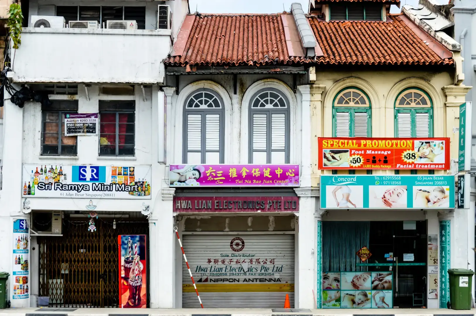 Row of shopfronts with white and pastel facades, arched windows, and colorful signboards on a street under a cloudy sky.
