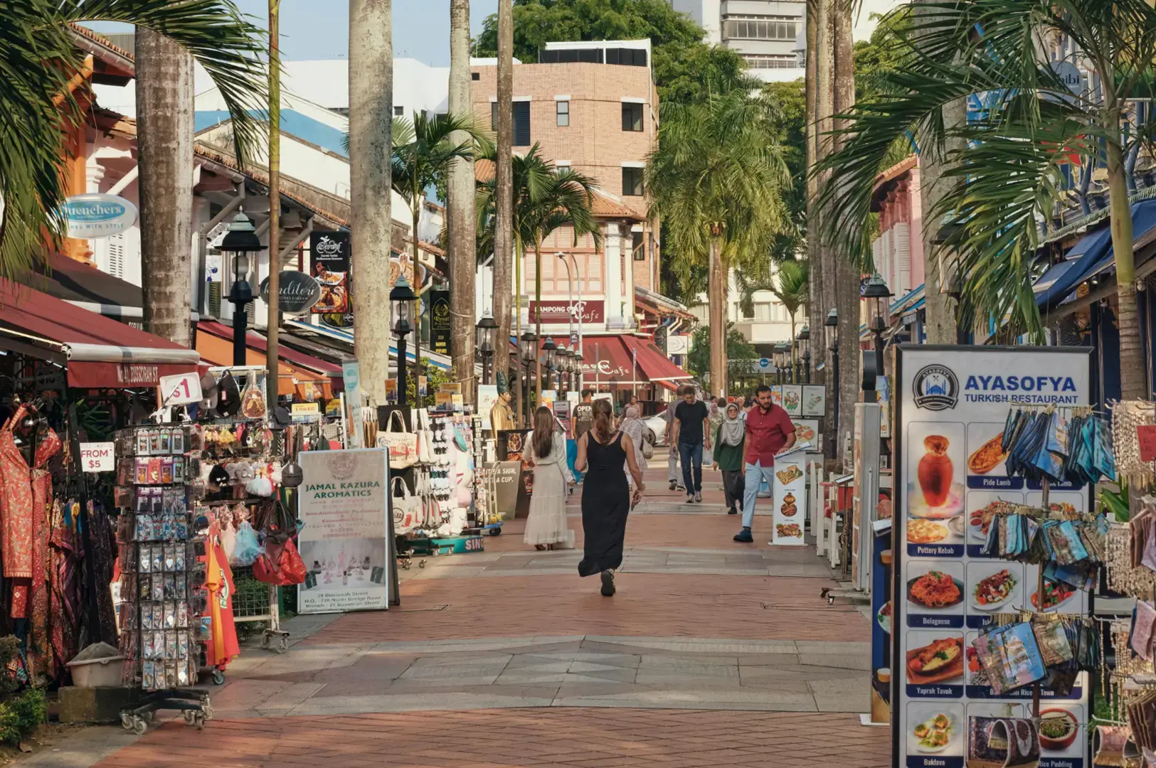 A sunny outdoor market street lined with tall palm trees and shops selling goods, with shoppers walking along the wide walkway between storefronts and signs.