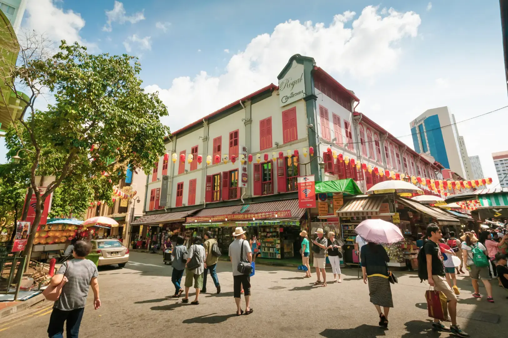 Sunny street scene with pedestrians and umbrellas, storefronts and colorful buildings lining both sides, and a prominent white building with red roof and signs in the center under a blue sky.
