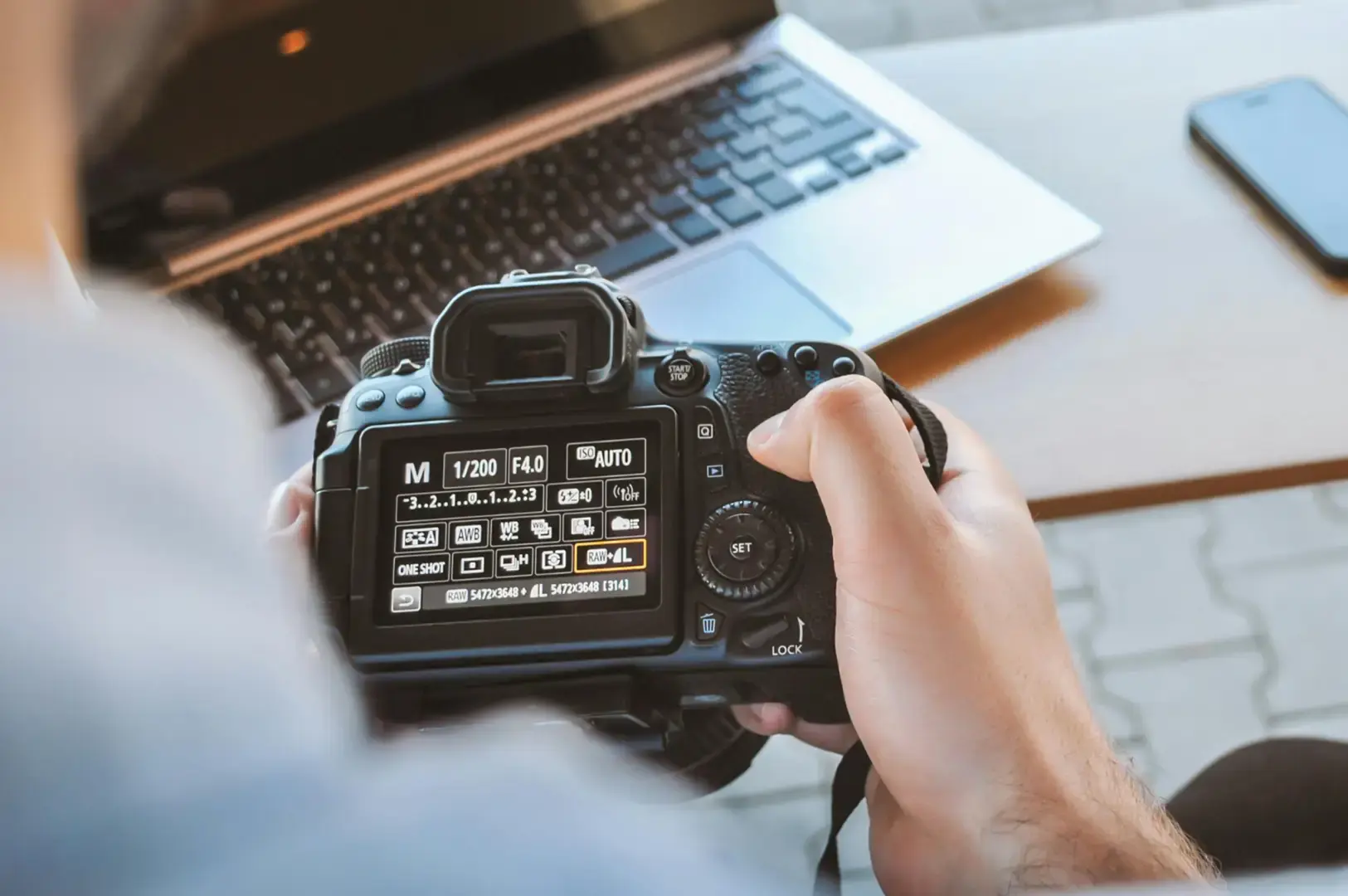 A person holding a DSLR camera with an LCD menu displayed, standing beside an open laptop and a smartphone on a desk, with a hand adjusting the camera controls.