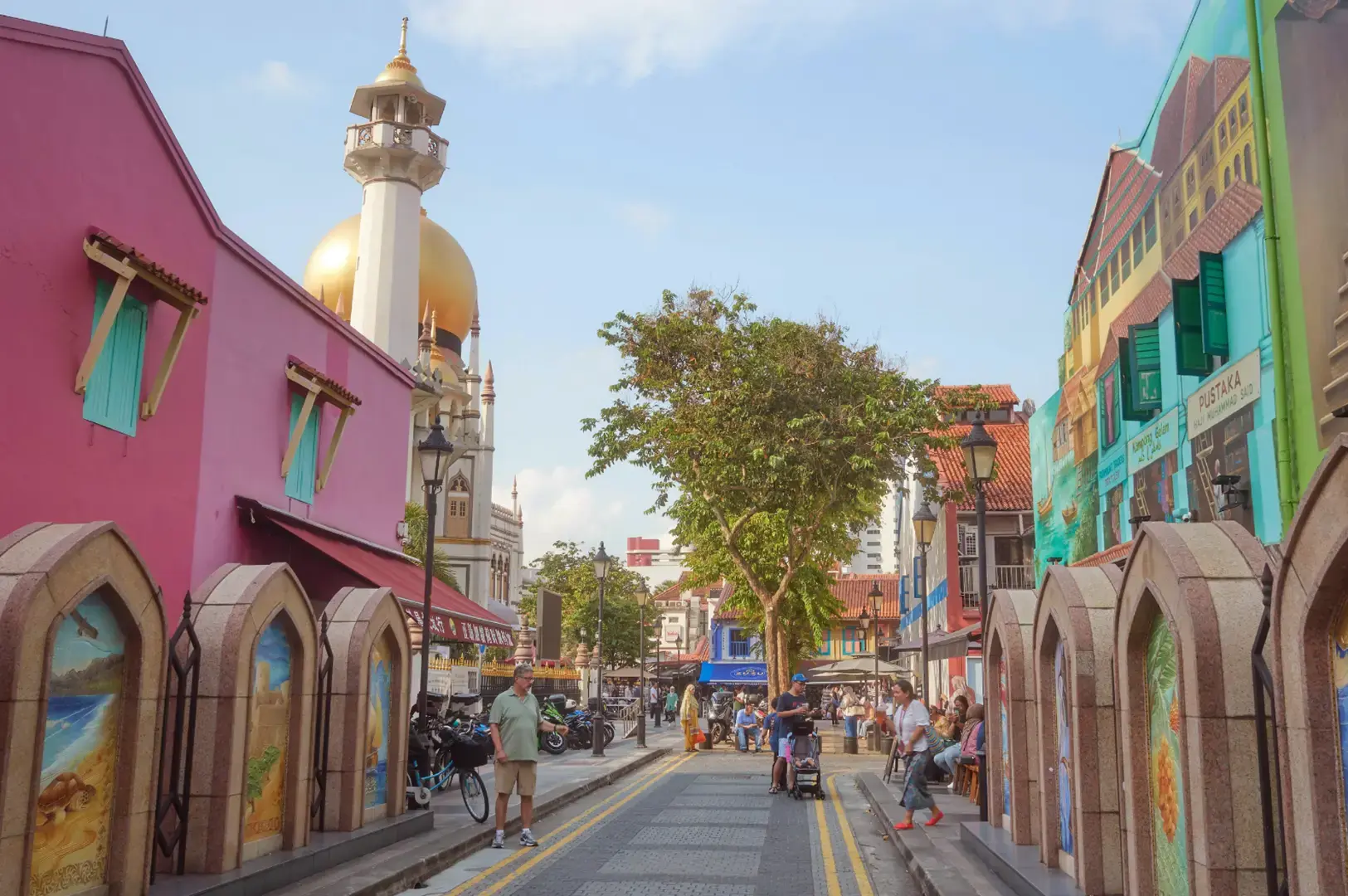A sunny pedestrian street with colorful buildings on both sides, arched murals along the walls, people walking, and a golden-topped tower in the distance under a blue sky.