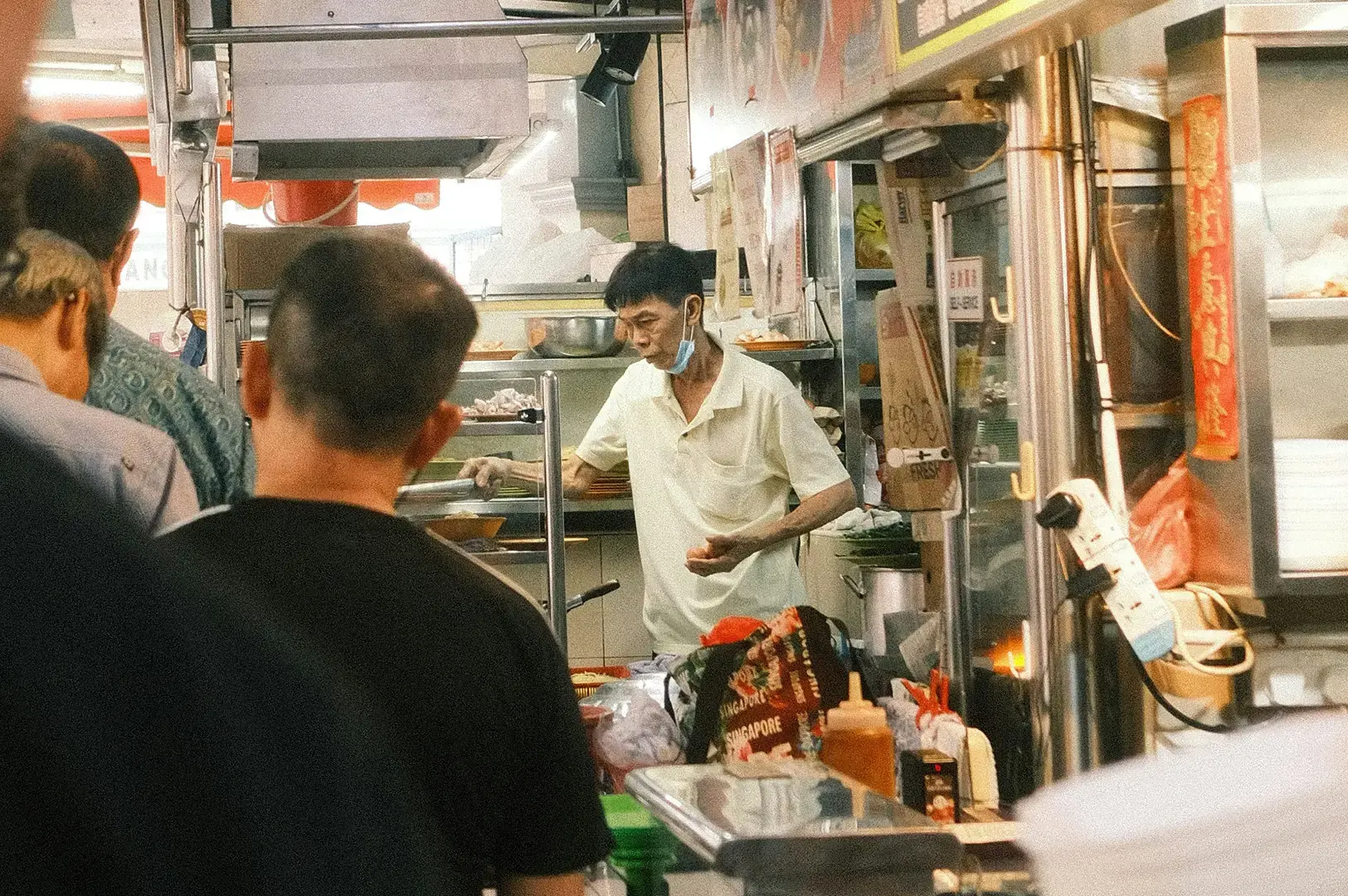 Older man preparing food at a busy Singapore hawker stall, with customers waiting in the foreground.