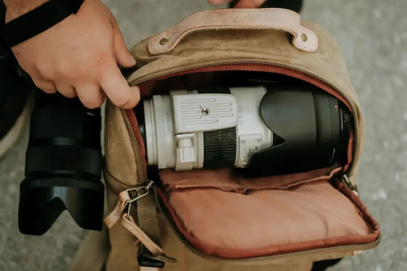 A close-up shows a beige camera case open on the ground, with a white-and-black lens and internal components visible as a hand reaches in to handle the gear.