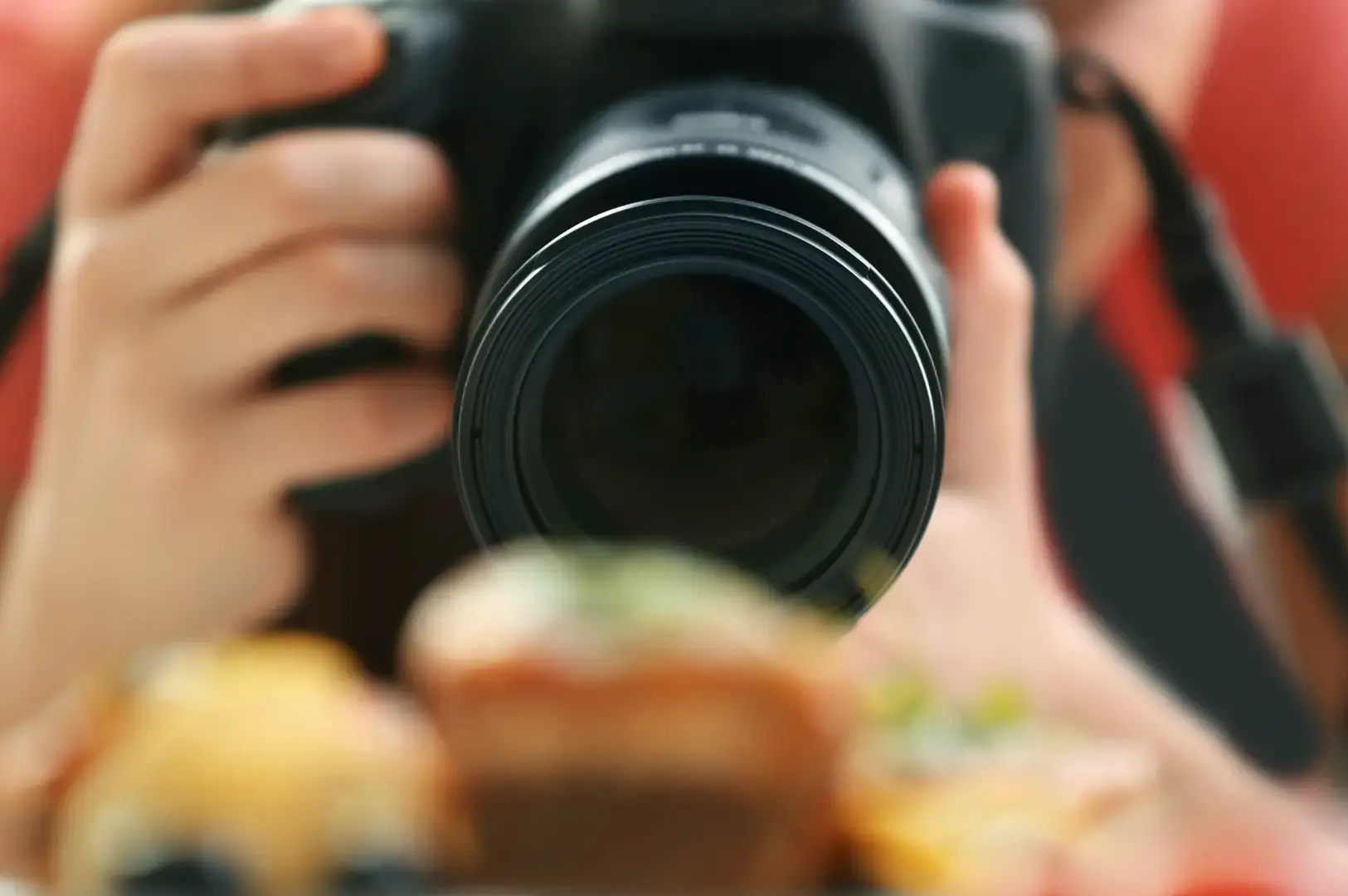 A person holding a black camera with a large lens to eye level, photographing a colorful table or scene in the foreground with a softly blurred background.