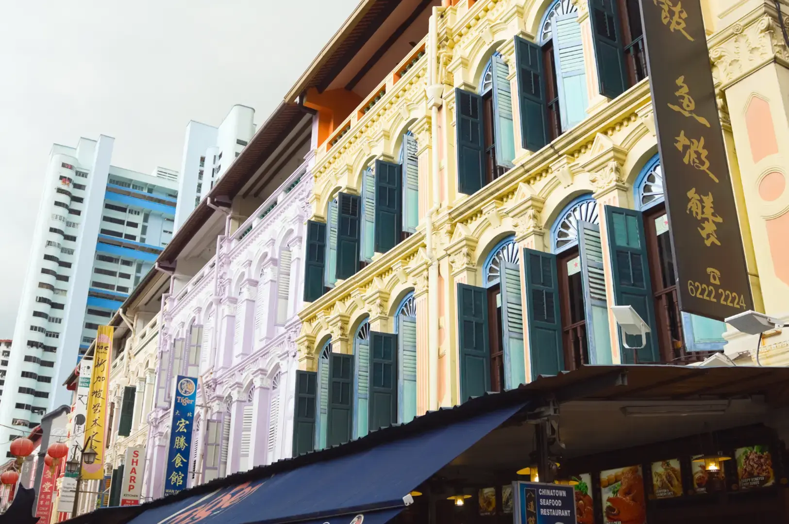Colorful historic shopfront along a street with ornate gold trim, arched windows, and vertical banners; a dark awning covers storefronts, with tall buildings in the background.